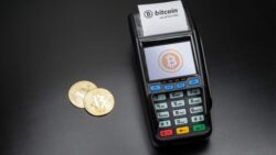 Card reader with a Bitcoin logo on the display and the note “Bitcoin accepted here,” next to two gold Bitcoin coins on a dark background – symbolizing cryptocurrencies as a payment method in retail.